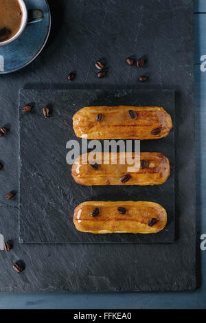 Coffe eclairs with cup of fresh black coffee and coffee beans on black slate board over blue wooden surface. With space for text Stock Photo