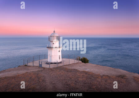 Beautiful old white lighthouse on the ocean coastline on the mountain at sunset. Summer seascape Stock Photo