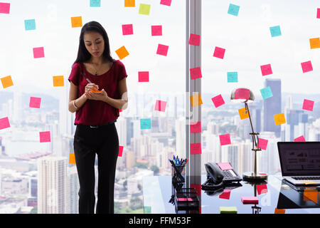 Young woman working as secretary in office, leaning on big skyscraper window. She writes adhesive notes with tasks on window. Stock Photo