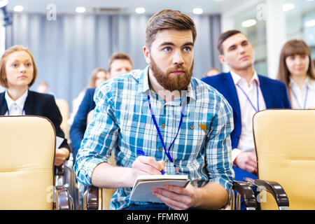 Focused serious young man listening and making notes on presentation in conference hall Stock Photo