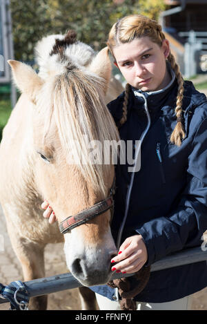 Young woman with braids holding the head of a Fjord horse on a farm on an sunny autumn day looking at camera Stock Photo