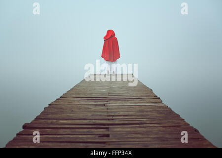 Red hooded woman looks at foggy lake on a wooden pier . Sadness and loneliness concept Stock Photo