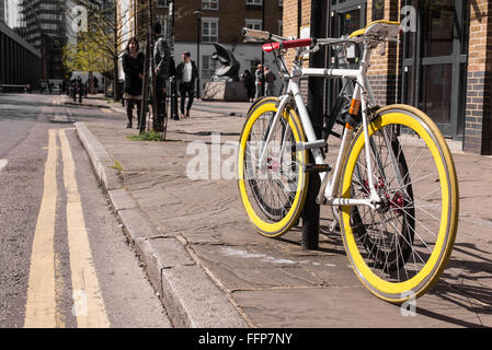 Modern single gear bicycle with yellow tyres locked to a street lamp. Blurred background with people walking in a street. Stock Photo