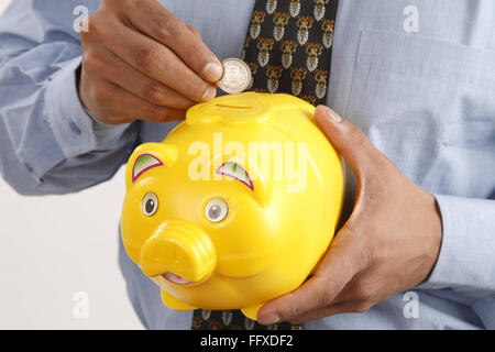 Businessman holding plastic piggy bank in one hand and inserting through slot five rupees coin MR#703T Stock Photo