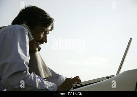 Indian businessman wearing light blue shirt and tie using laptop on bonnet of car and talking on mobile phone Pune Maharashtra Stock Photo