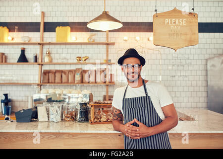 Portrait of happy young man wearing an apron and hat leaning to a cafe counter. Male barista standing at coffee shop looking at Stock Photo