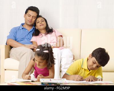 Children doing drawing work parents resting on sofa behind them in house MR#702R,MR#702S,MR#702T,MR#702U Stock Photo