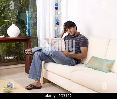 Young man thinking holding newspaper sitting on sofa MR#702V Stock Photo