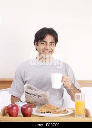 Young man holding newspaper and mug having breakfast on bed MR#702V Stock Photo