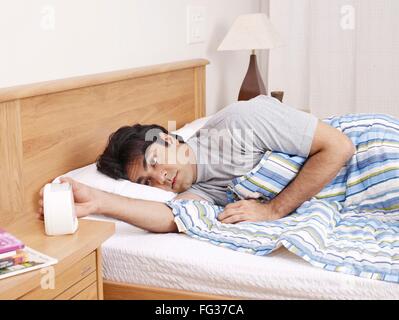 Young man laying on bed hand stopping ringing alarm clock MR#702V Stock Photo