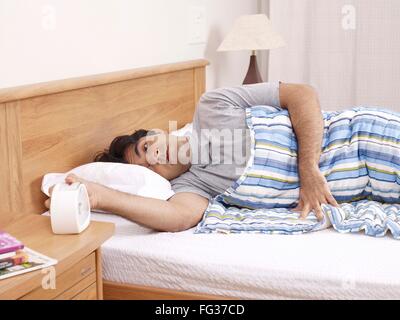 Young man laying on bed hand stopping ringing alarm clock MR#702V Stock Photo