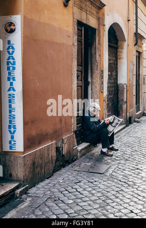 An elederly man reads a newspaper while sitting on a door step in Rome, Italy Stock Photo