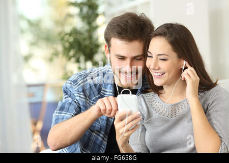 Happy couple sharing music from smart phone sitting on a couch at home Stock Photo