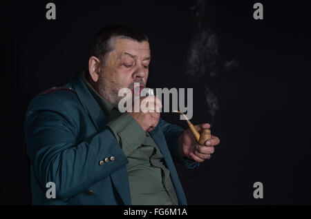 Portrait of an old retired military man who is ready to drink glass of vodka while smoking tobacco-pipe Stock Photo