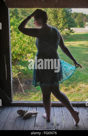 A Red-Headed, Caucasian Teenage Girl Wearing A Turquoise Dress And No Shoes Stands In The Hay Loft Of An Old Barn Framed By The Open Loft Window Ov... Stock Photo