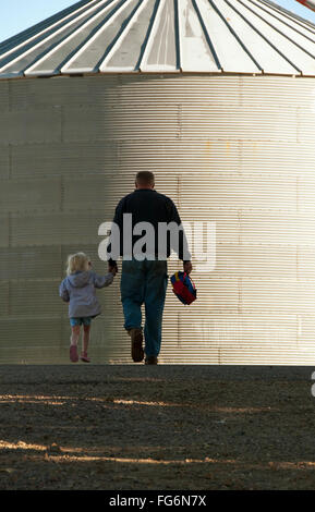 Agriculture - A farmer and his young daughter walk hand-in-hand through his farm yard with a grain bin in the background / near Sioux City, Iowa, USA. Stock Photo