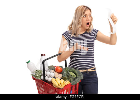 Shocked woman holding a shopping basket full of groceries and looking at the bill in disbelief isolated on white background Stock Photo