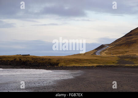 A black beach on the foreground, and an empty road behind  surrounded by yellow fields and with a dark blue sky as a background Stock Photo