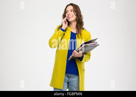 Happy lovely young woman with folders standing and talking on mobile phone over white background Stock Photo