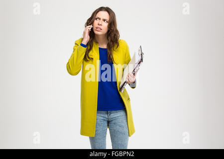 Thoughtful frowning young woman holding clipboard and using smartphone over white background Stock Photo
