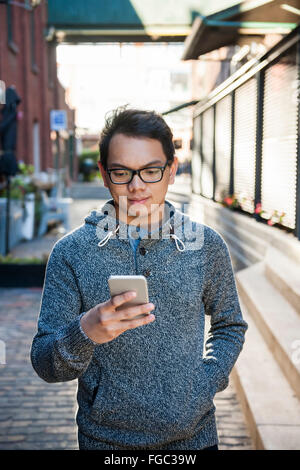 Young asian man wearing glasses looking into smartphone while walking on city street Stock Photo