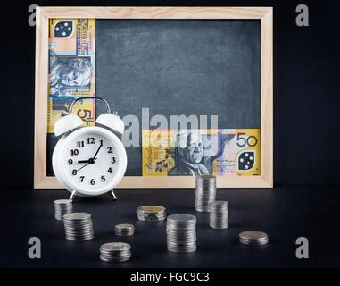 Vintage alarm clock showing 9 o'clock, coin towers and 50 Australian dollars bills on empty chalkboard. Time is money business c Stock Photo