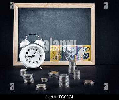Vintage alarm clock showing 9 o'clock, coin towers and 50 Australian dollars bill on empty chalkboard. Time is money business co Stock Photo