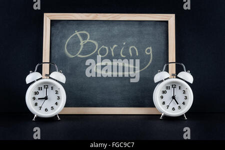 Two vintage alarm clocks showing 9 and 5 o'clock with the word "boring" handwritten on chalkboard on black background. Nine to f Stock Photo