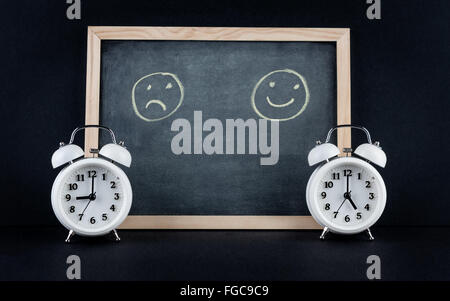 Two vintage alarm clocks showing 9 and 5 o'clock with happy and sad smileys on chalkboard on black background. Nine to five corp Stock Photo