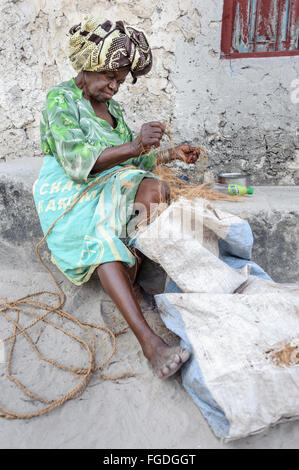 Zanzibari old woman with colourful attire making a rope with coconut fibers outside her home. Stock Photo