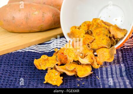 A bowl of homemade baked sweet potato chips poured over a kitchen towel and raw vegetable on a cutting board. A healthy sweetpot Stock Photo