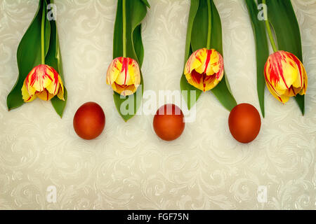 Three Easter eggs, colored red and yellow tulips with green leaves on a light background. The view from the top. Stock Photo