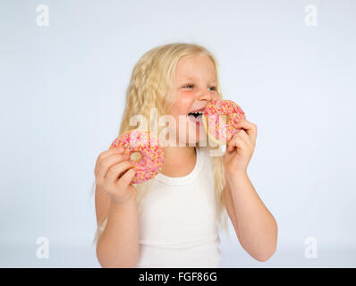 Young girl with long blonde hair holding two pink iced donuts, laughing Stock Photo