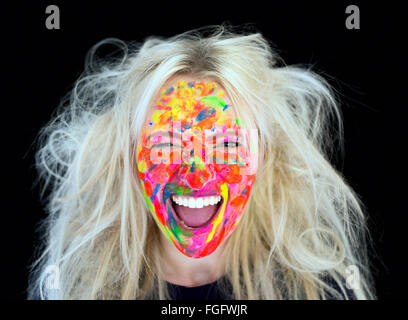 Woman with messy blonde hair with face covered in multi coloured paint laughing Stock Photo