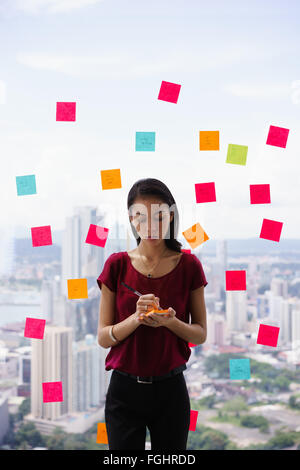 Young woman working as secretary in office, leaning on big skyscraper window. She writes adhesive notes with tasks on window. Stock Photo