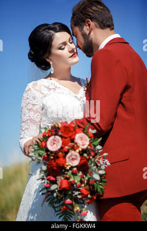 Bride and groom, lovely couple outdoor, wedding bridal bouquet with red flowers. Blue sky, green grass in a background. Stock Photo
