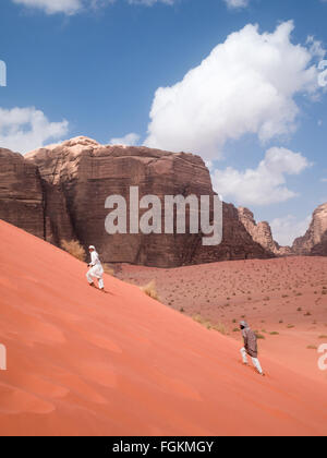 Two men dressed in Arabian costume climb up the red sand dunes of the Wadi Rum desert Stock Photo