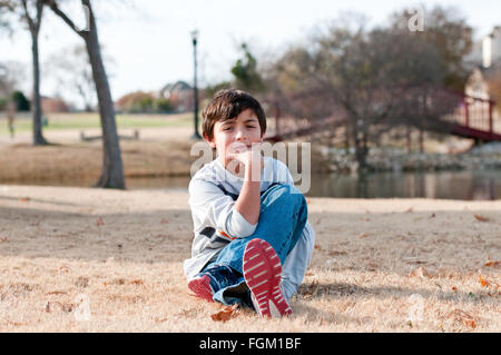 Cute little boy sitting in grass during the fall at a park with pond and bridge in background. Stock Photo