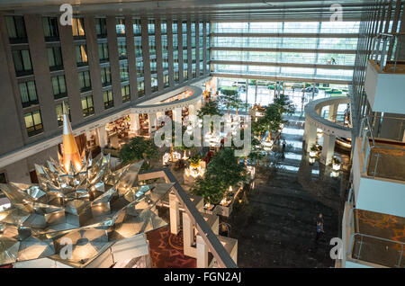 Novotel Suvarnabhumi Airport Hotel Modern Atrium Bangkok Thailand | BANGKOK, Thailand — The opulent interior of the Novotel Suvarnabhumi Airport hotel in Bangkok features a modern atrium where contemporary design meets traditional Thai elements. The spacious, multi-story lobby is characterized by a striking central sculpture, lush indoor trees, and expansive glass walls, creating a luxurious and comfortable environment. Located directly opposite Suvarnabhumi Airport (BKK), the hotel offers convenient access for international travelers. Its strategic position and comprehensive facilities make i Stock Photo
