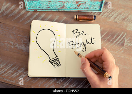 Retro effect and toned image of a woman hand drawing a light bulb next to BE BRIGHT text with a fountain pen on a notebook. Copy space available, ideas creativity conceptual image Stock Photo