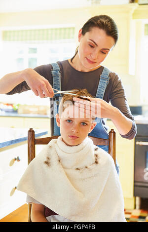 Portrait unhappy boy getting haircut from mother in kitchen Stock Photo