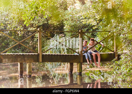 Brother and sister sitting on footbridge in park with trees Stock Photo