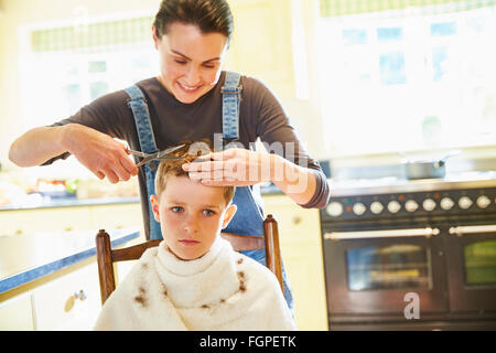 Unhappy boy getting haircut from mother in kitchen Stock Photo
