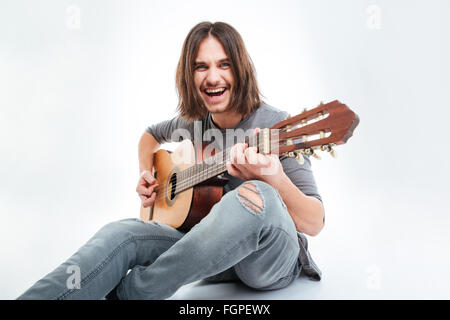 Cheerful handsome young man with long hair sitting and playing guitar over white background Stock Photo