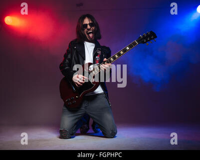 Crazy young guitarist with long hair playing electric guitar on his knees and shoing tongue over colorful smoky background Stock Photo