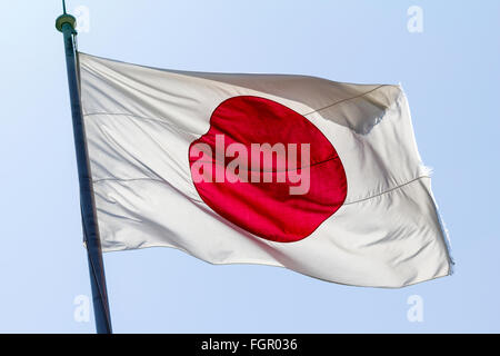 Japanese rising sun flag, red ball on white background, fluttering in breeze against clear blue sky. Stock Photo