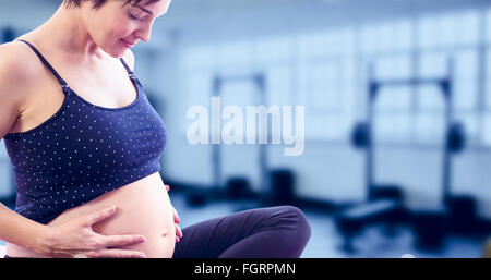 Composite image of happy pregnant woman holding belly while sitting on exercise mat Stock Photo