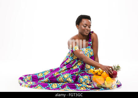 Smiling charming african american young woman in bright sundress sitting with glass bowl of fruits over white background Stock Photo