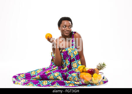 Cute lovely pensive african american young woman in colorful sundress sitting and holding orange over white background Stock Photo