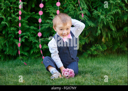 little boy in a suit with a bouquet sitting on the grass Stock Photo